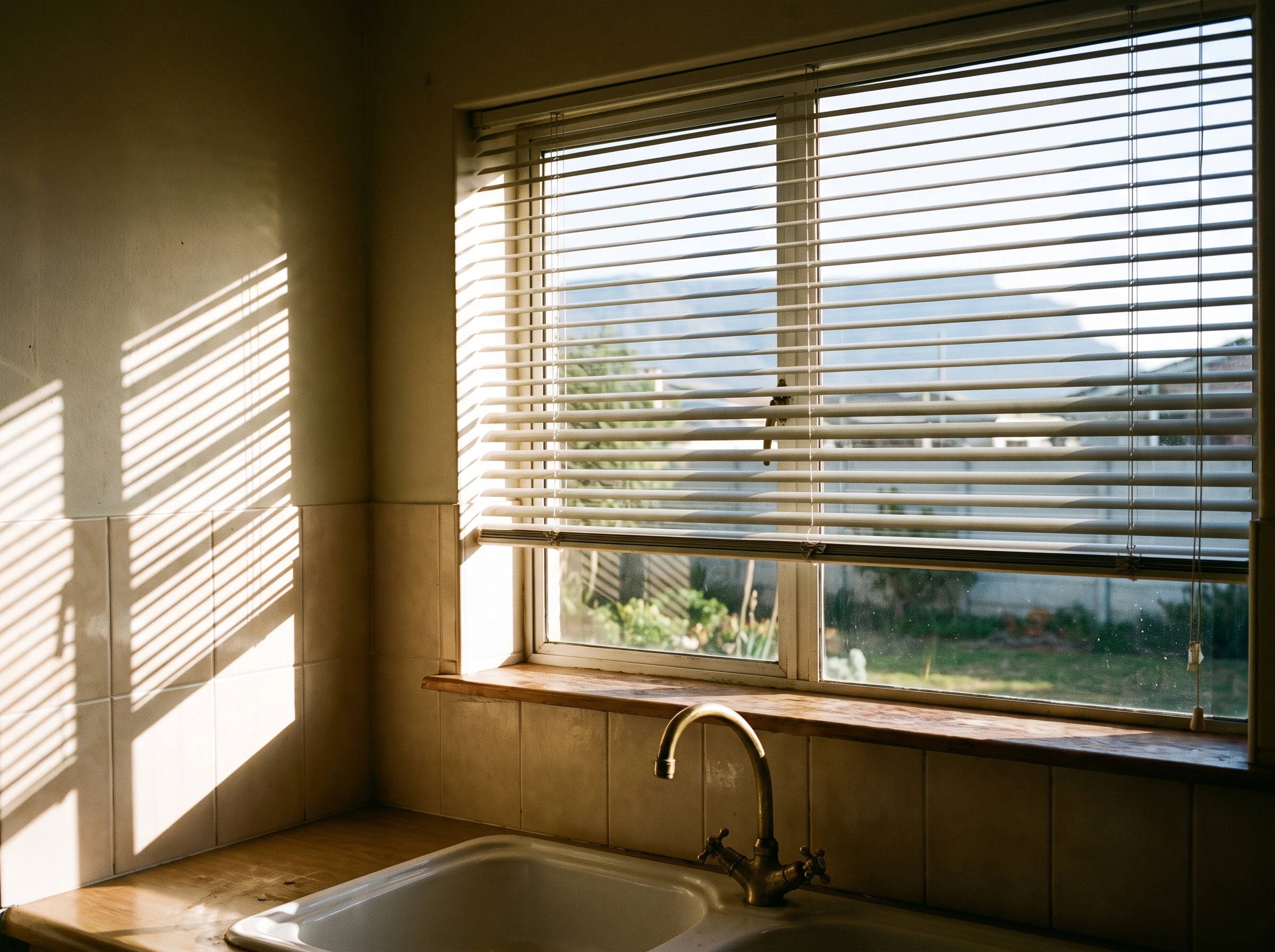 Aluminium venetian blind with tilted slats above a kitchen sink window in a Kraaifontein family home, morning light striping through onto the splashback